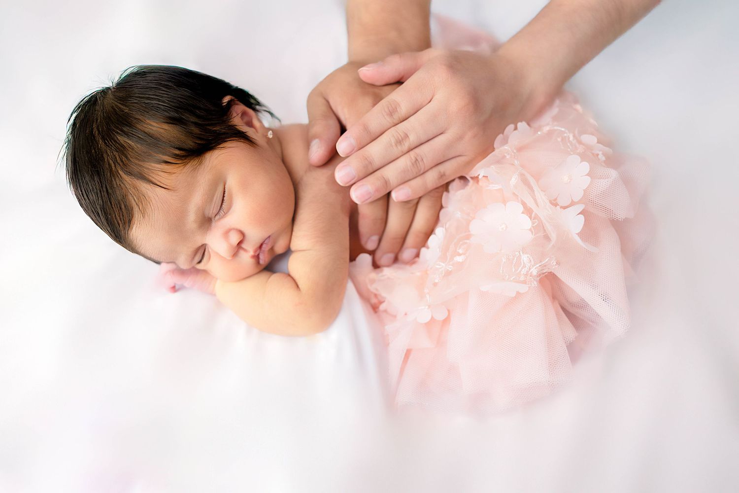 Sleeping baby wearing a soft pink tulle dress lies peacefully on white fabric while hands gently rest nearby. #ArtesiaPhotographer