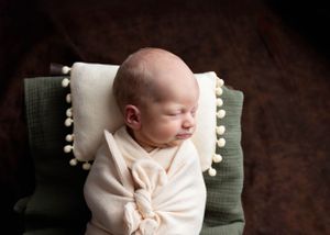 A sleeping newborn in a cozy cream-colored wrap rests on a green chair with white pom-pom trim pillow #RoswellNM #newbornphotographer