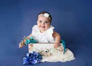 Baby wearing white dress sits in decorative box with blue hydrangeas against royal blue background. #CarlsbadNM #Carlsbadphotographer
