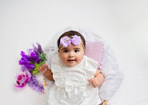 A baby in a white lace dress and floral headband lies on white fabric with purple flowers beside them. #NewbornPictures #Photographer #NM