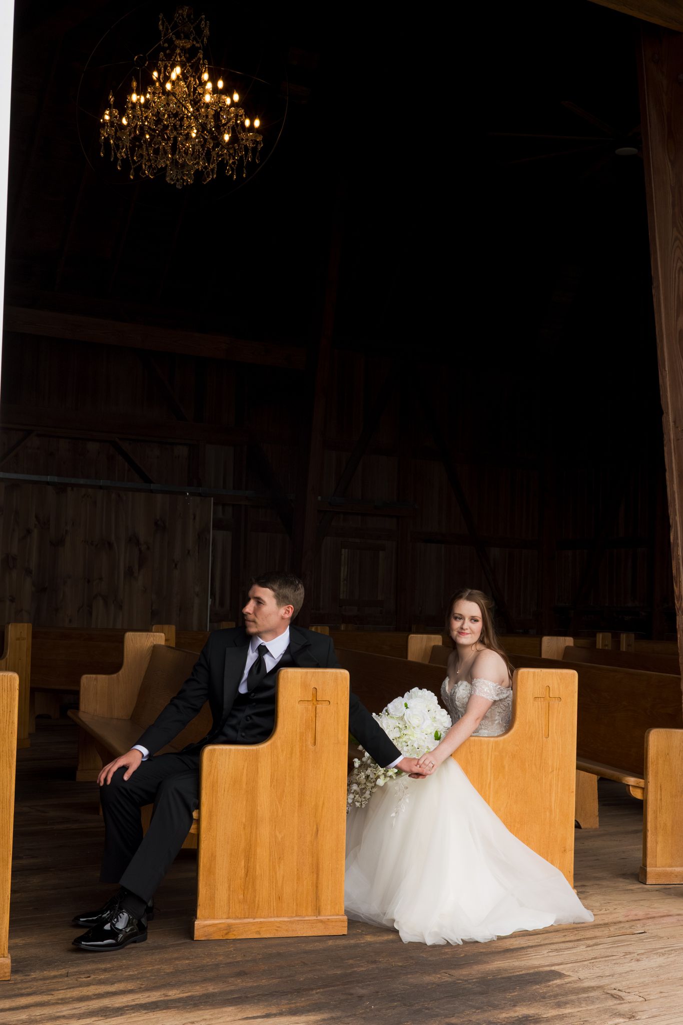 Wedding couple sitting apart on wooden church pews in a dimly lit room with chandeliers overhead.