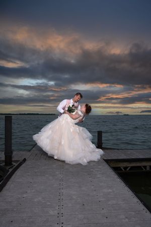 A romantic sunset photo of a couple posing on a waterfront dock in formal wedding attire.