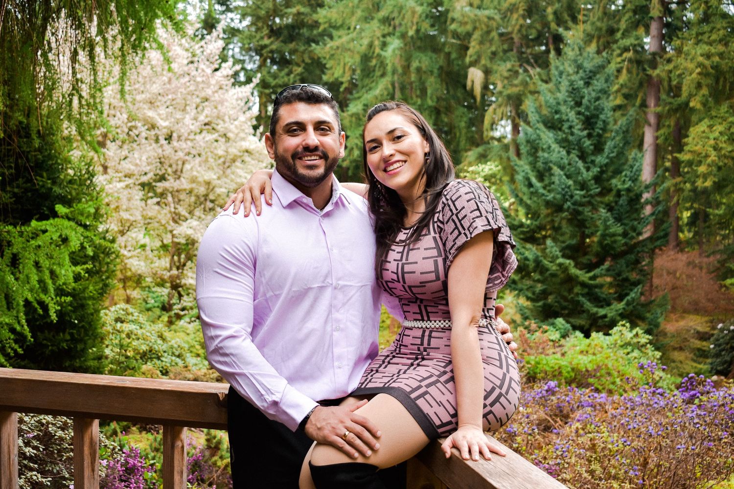 Couple sitting on a wooden bridge surrounded by lush greenery and flowers in a serene botanical garden setting.