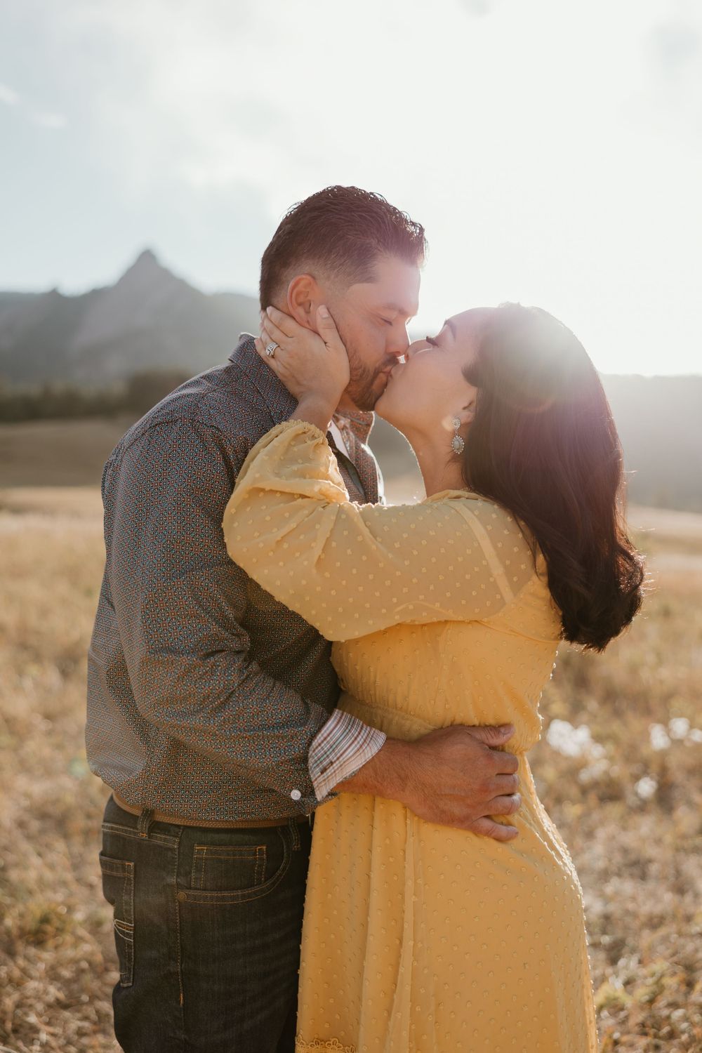 Romantic couple sharing intimate kiss in golden sunlit field with mountains in background.