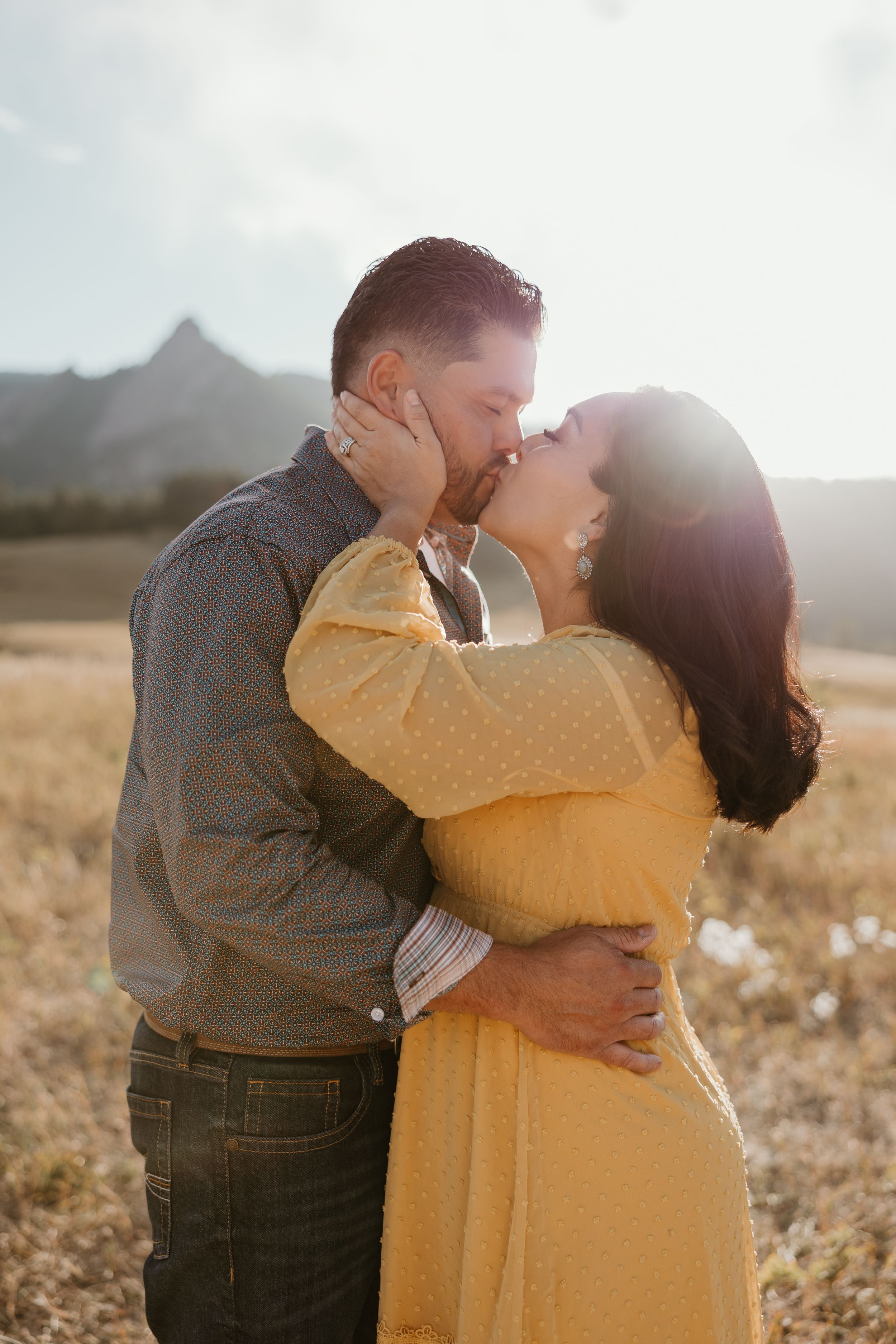 Romantic couple sharing intimate kiss in golden sunlit field with mountains in background.