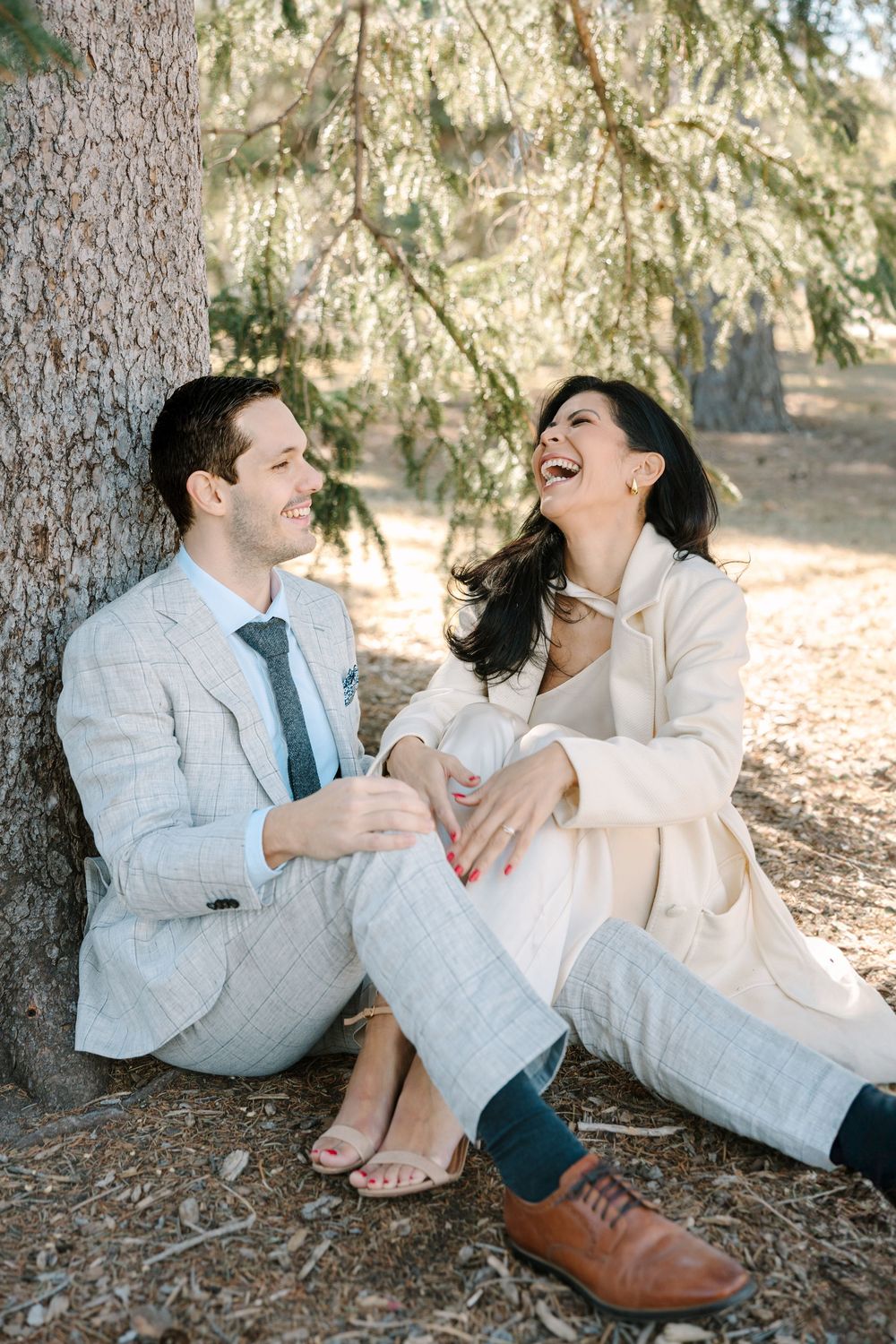 A couple laughs together while sitting under a pine tree.