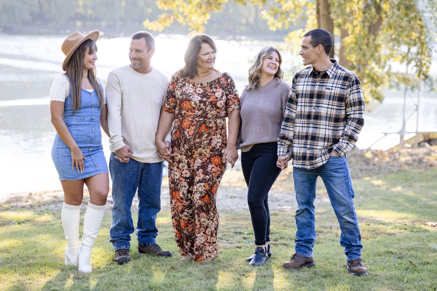 5 adults hold hands and smile and laugh together in front of a lake