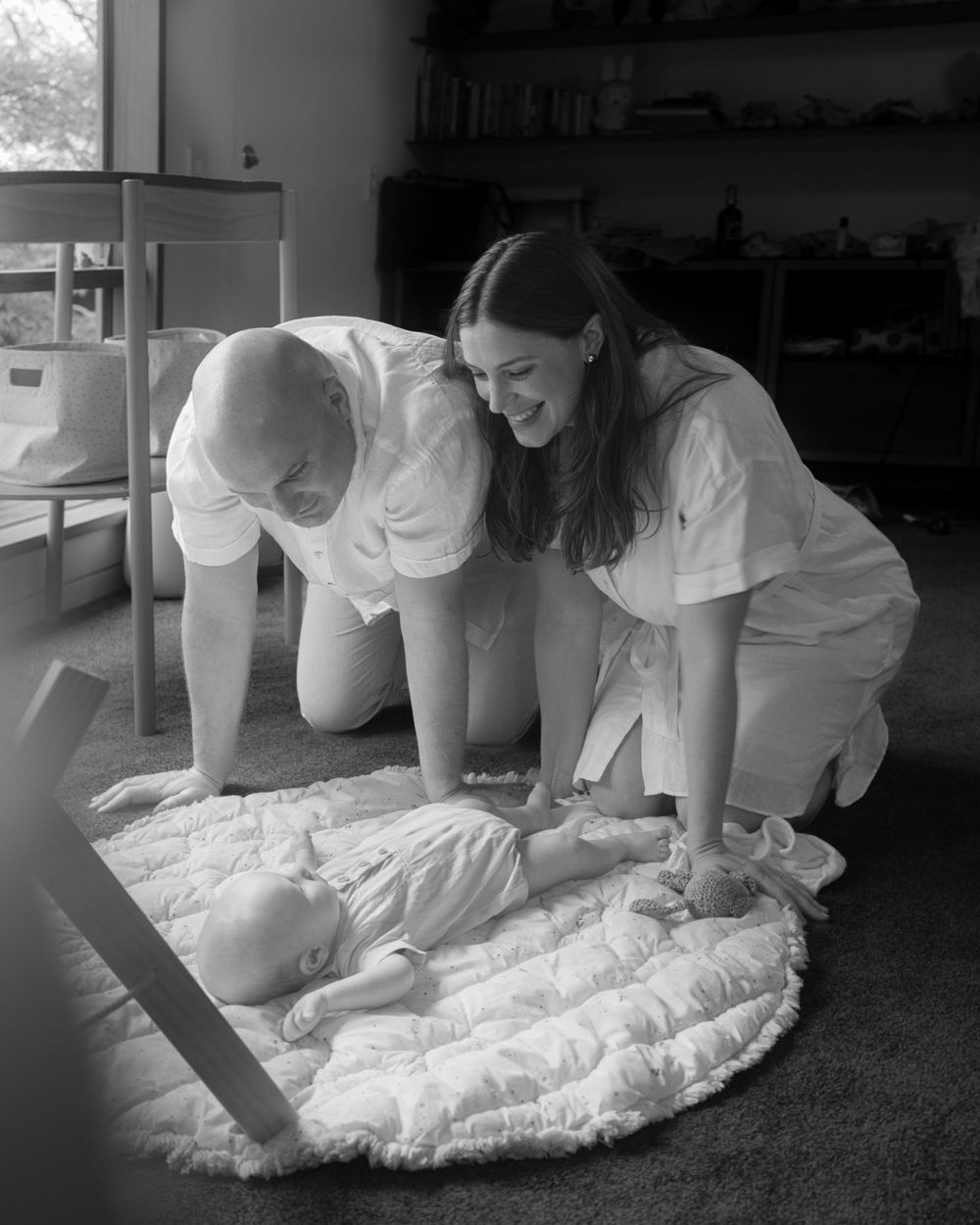 Parents lean down to interact with baby lying on quilted mat in home setting.