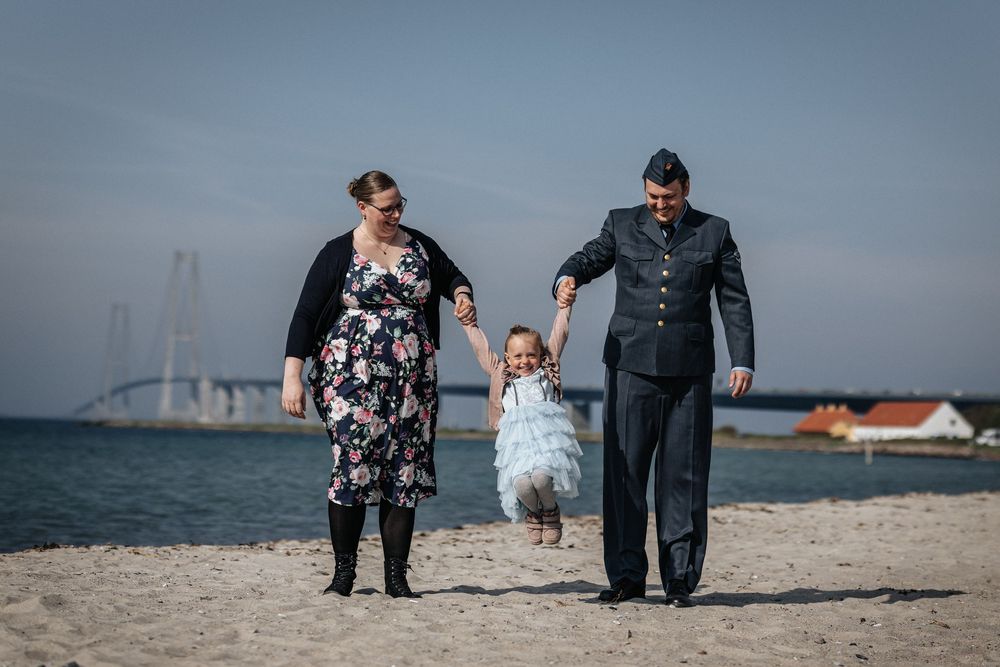 Three people walking together hand in hand on a beach with bridge and boats visible in the distant background.