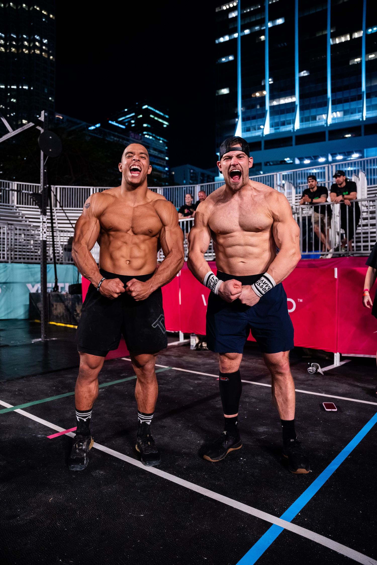 Two muscular athletes pose together at night with city lights in background.