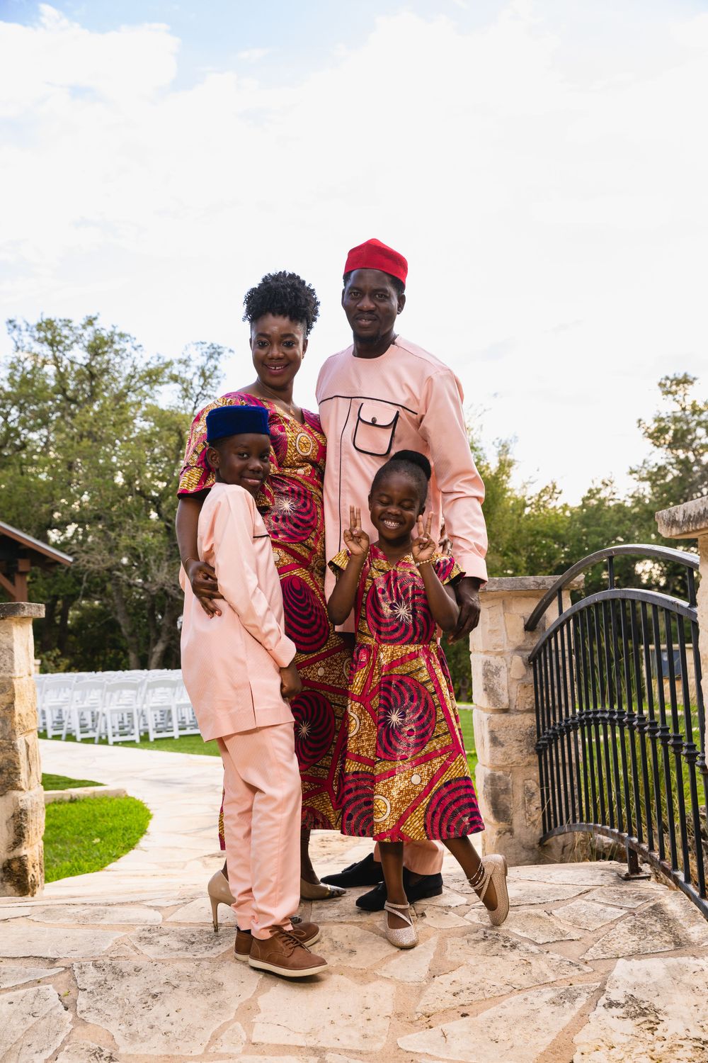 A family wearing coordinated pink and traditional African print outfits poses by a stone gate entrance.