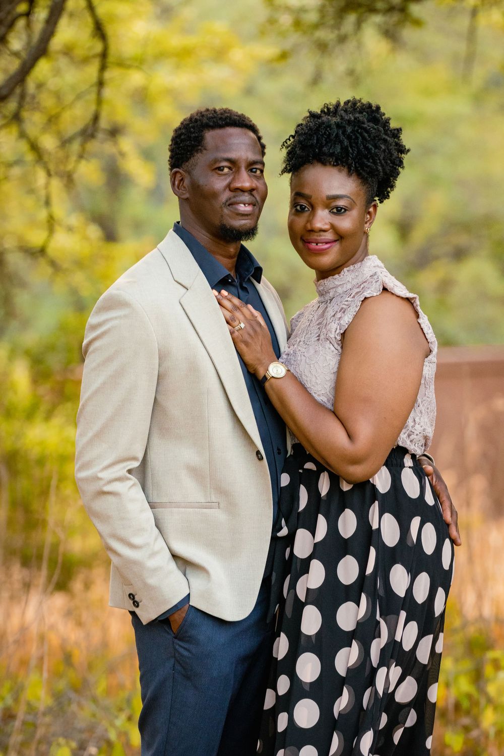 A couple poses together outdoors wearing a beige blazer and polka dot dress against fall foliage.