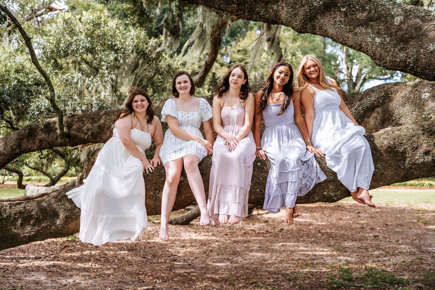 A group in white outfits poses together under a large sprawling tree branch in an outdoor setting.