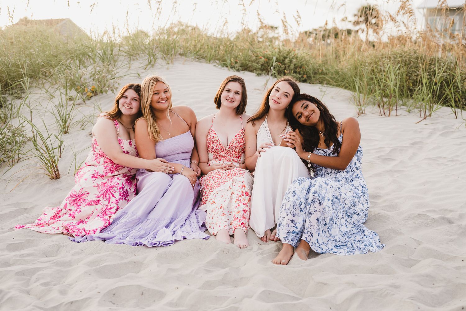 A group in floral and pastel dresses sits together on a sandy beach during sunset.