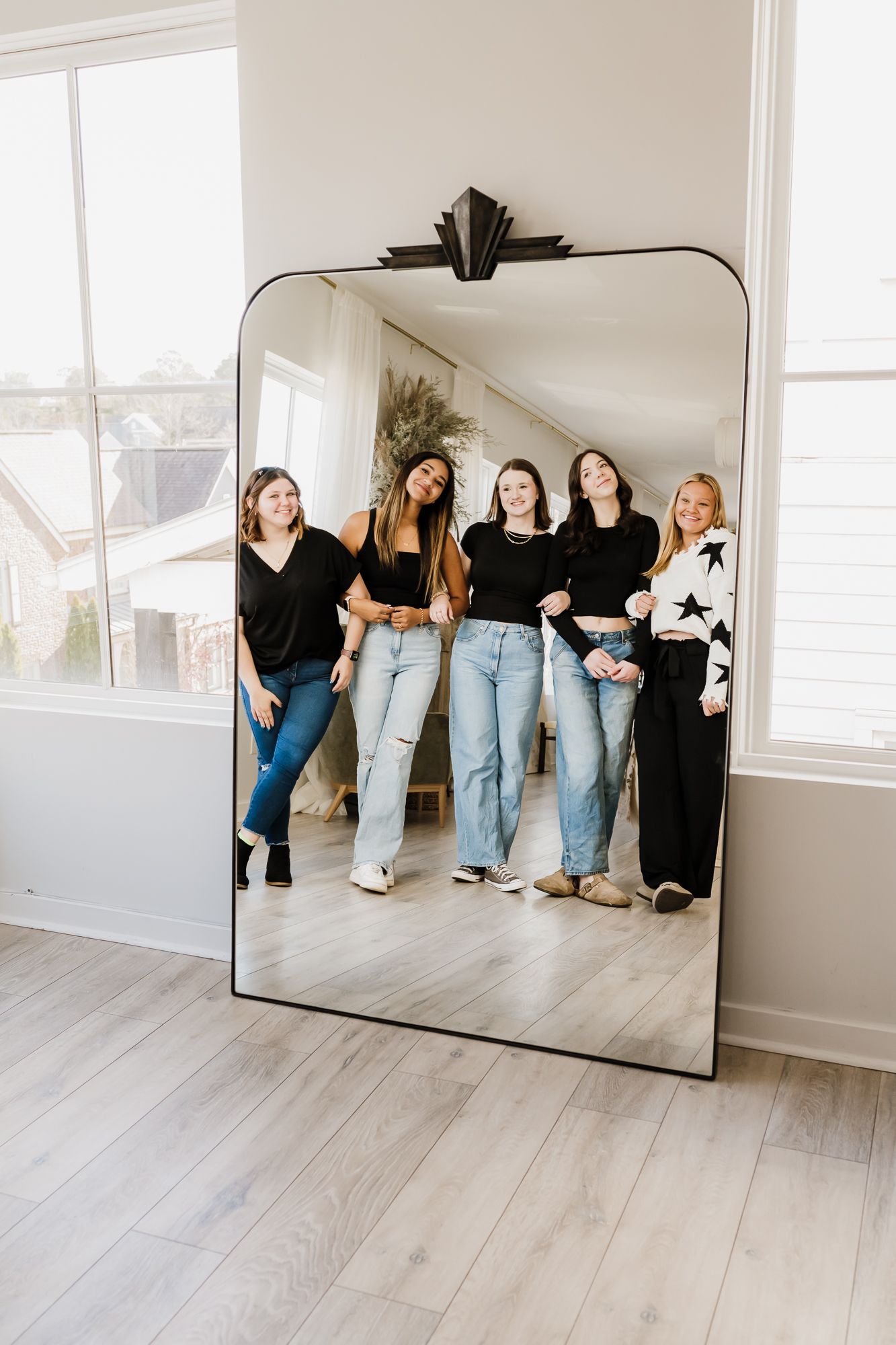 A group wearing matching black tops and jeans poses casually in a bright studio space with light wood flooring and white walls.