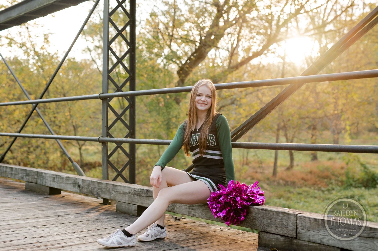 Cheerleader poses on rustic bridge with pink pom poms during sunset with fall colors in background.