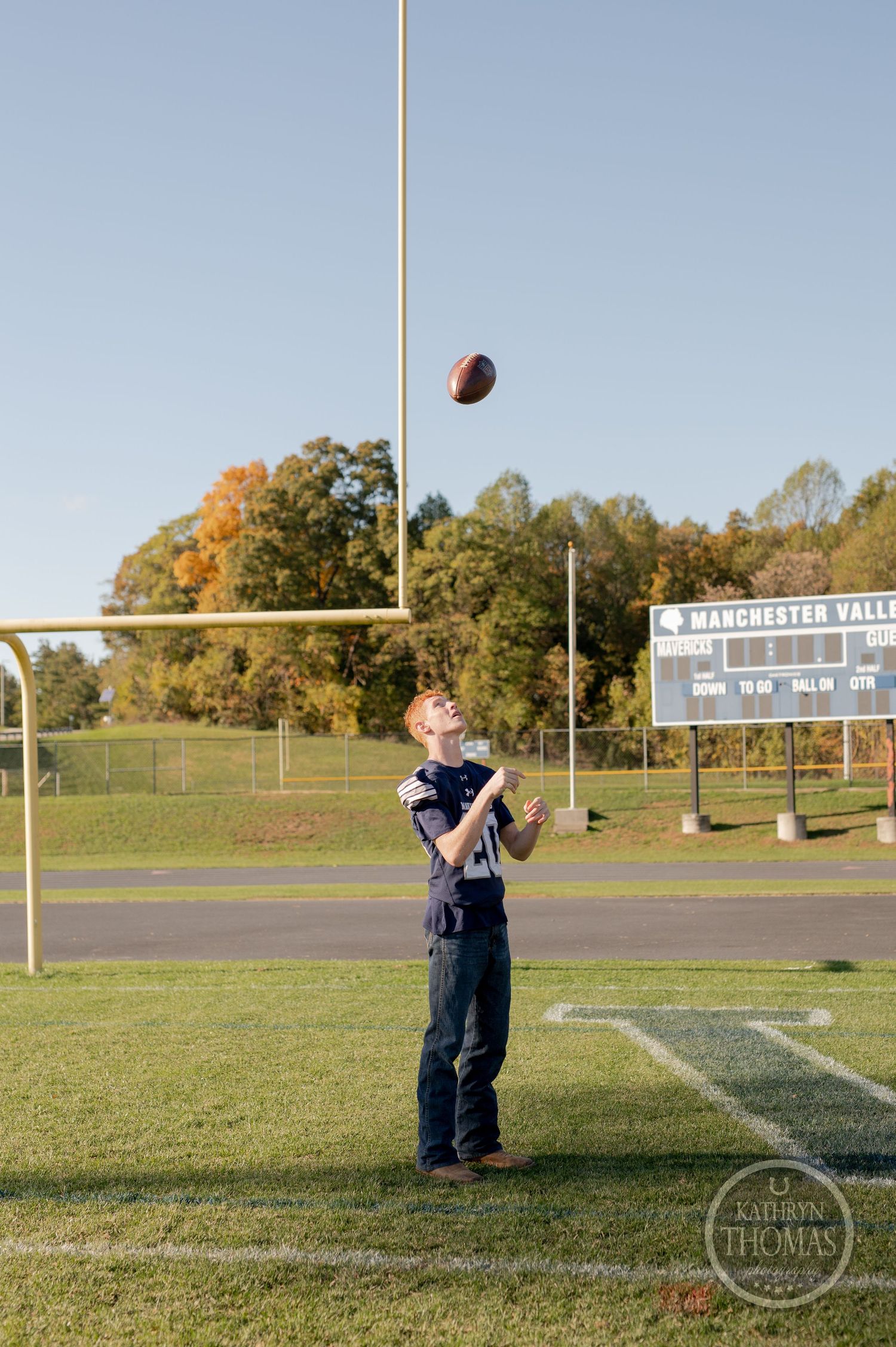Player throws football on field with goal post and scoreboard in background.