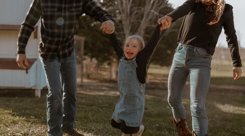 A small child holds hands with adults while jumping on a grassy lawn at sunset.