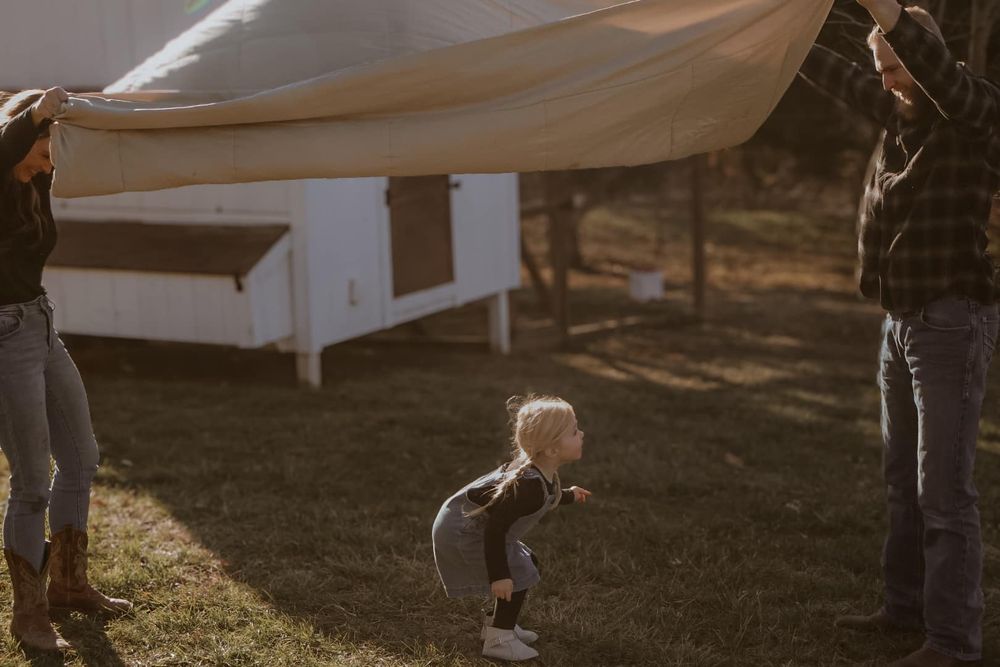A child plays under a white sheet being held up in a backyard.