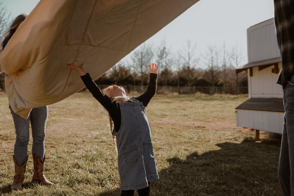 A child reaches up towards a white sheet being held overhead in golden sunlight.