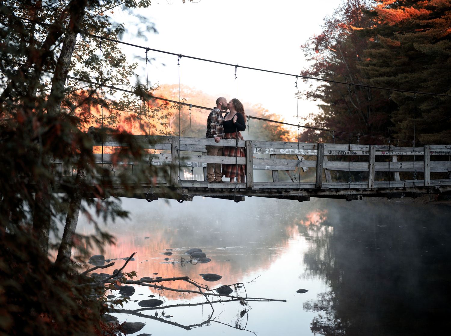 Panoramic view of a misty lake at sunset with trees and a bridge in the distance.
