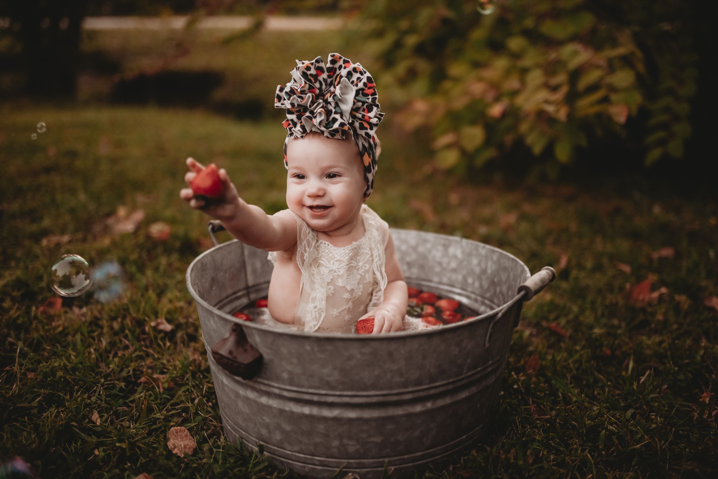 A child sits in a rustic metal tub surrounded by fall foliage and red apples.