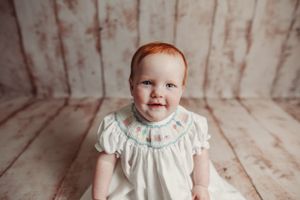 Baby in white dress during professional portrait session against distressed wooden backdrop.