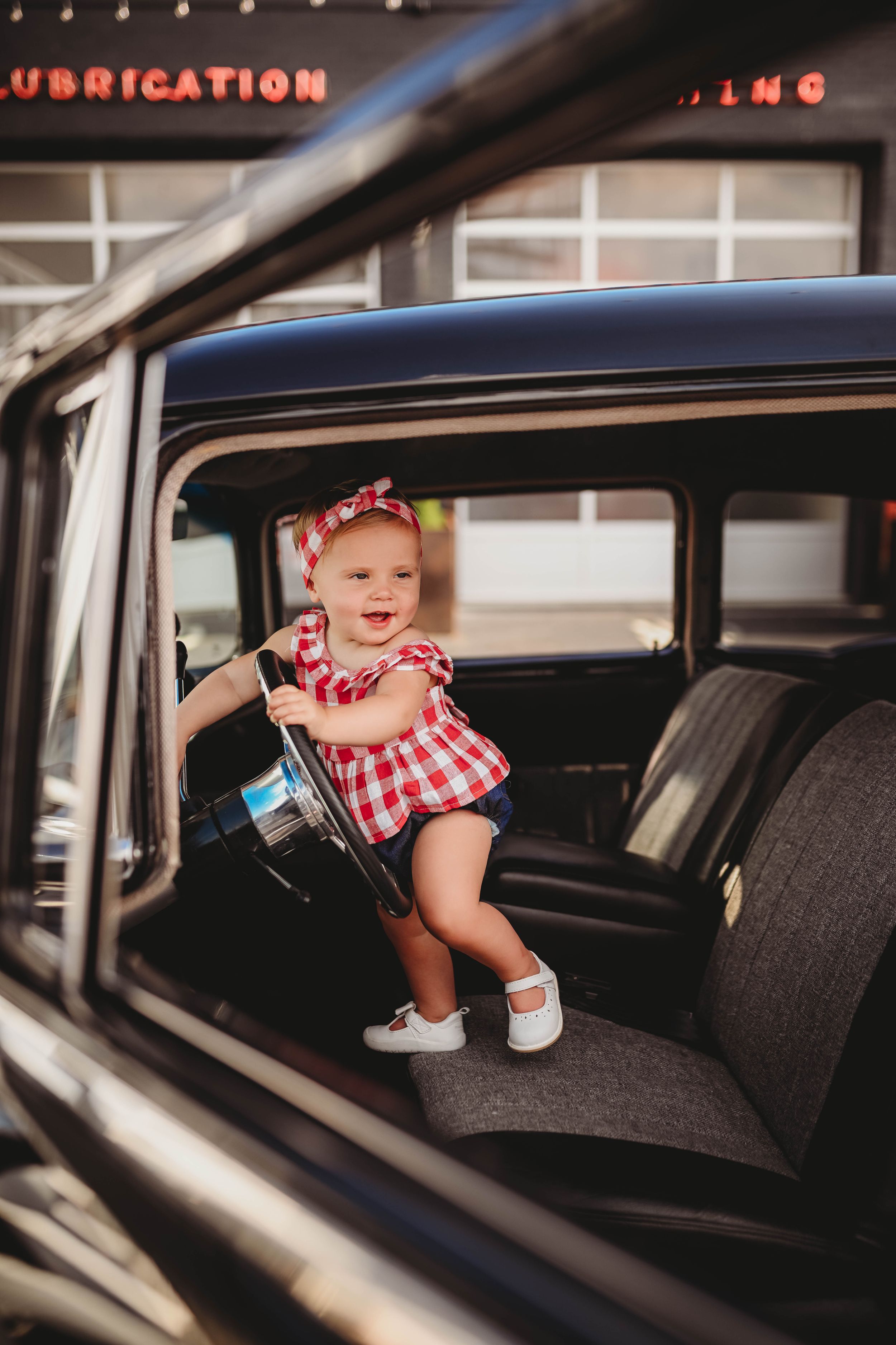 Baby in red gingham dress sitting inside vintage black car.