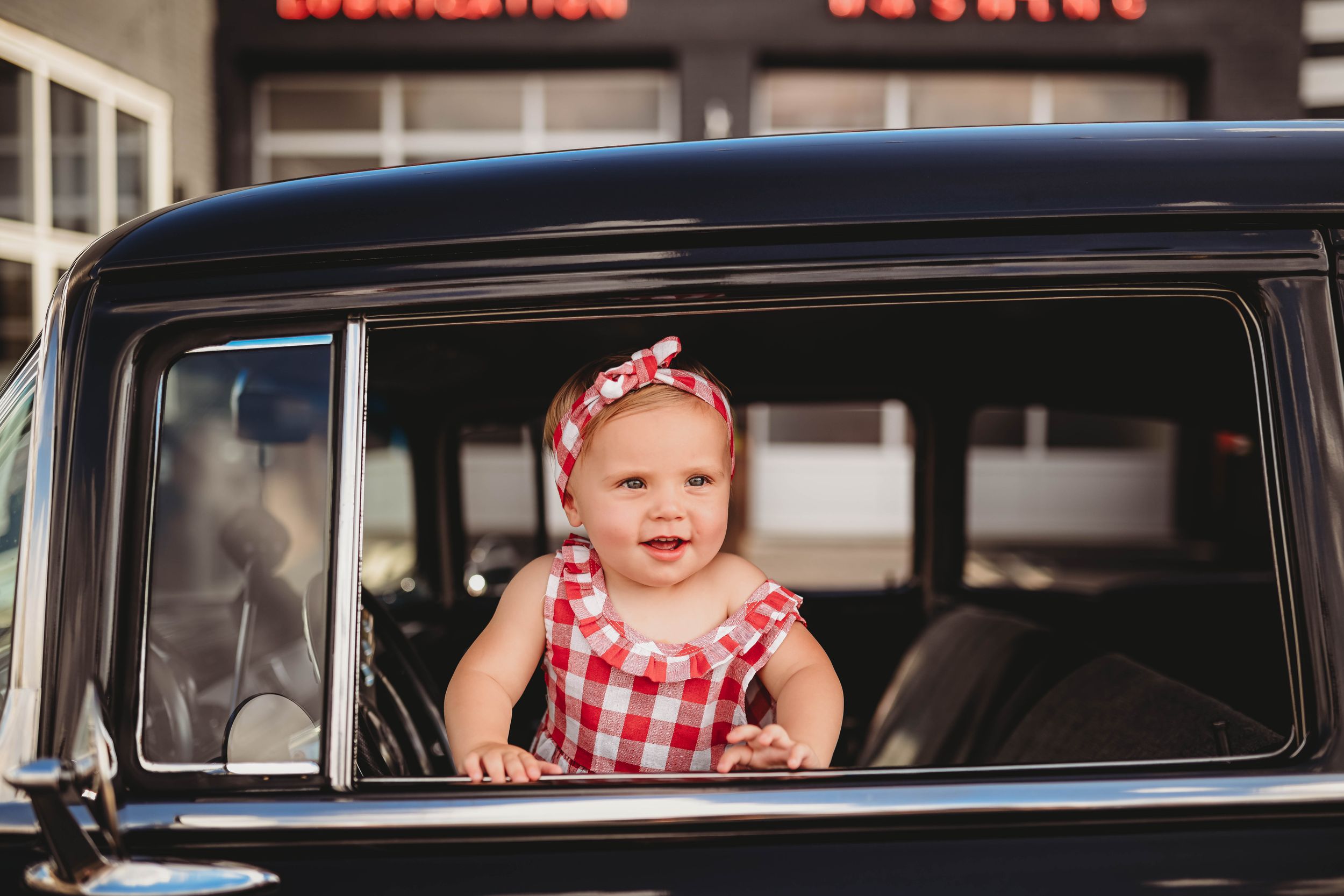 Baby in red checkered dress peering through classic car window.