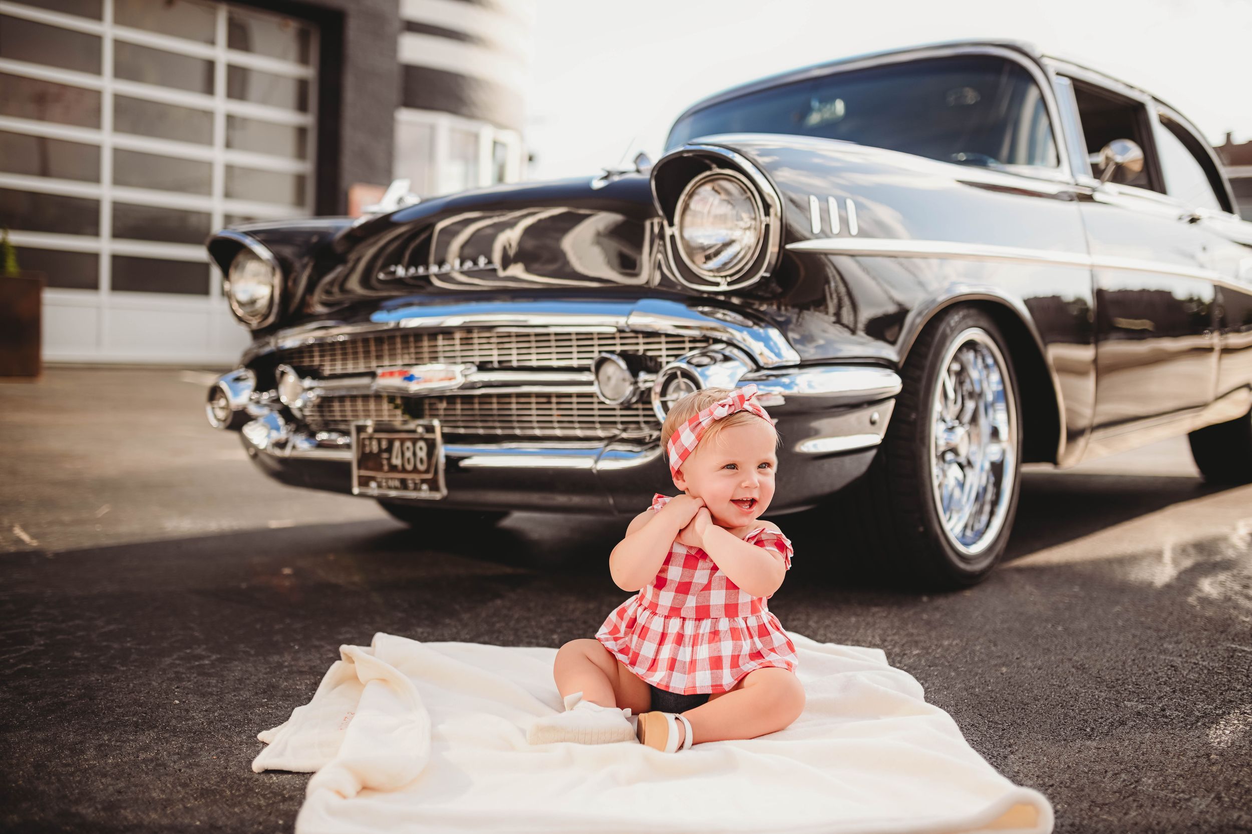 Baby in red dress posing with classic vintage automobiles.