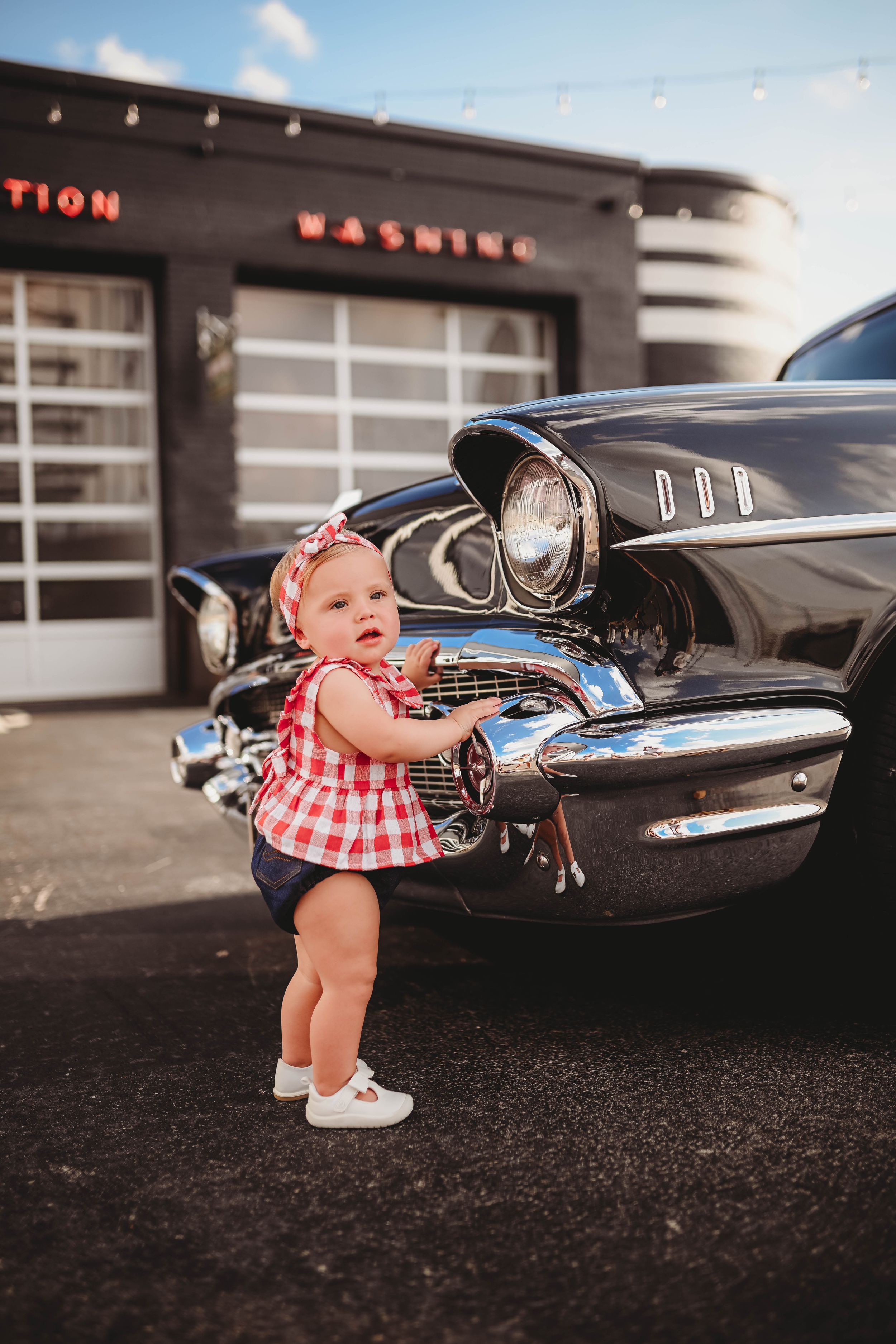 Baby in red dress standing next to classic black car outdoors.