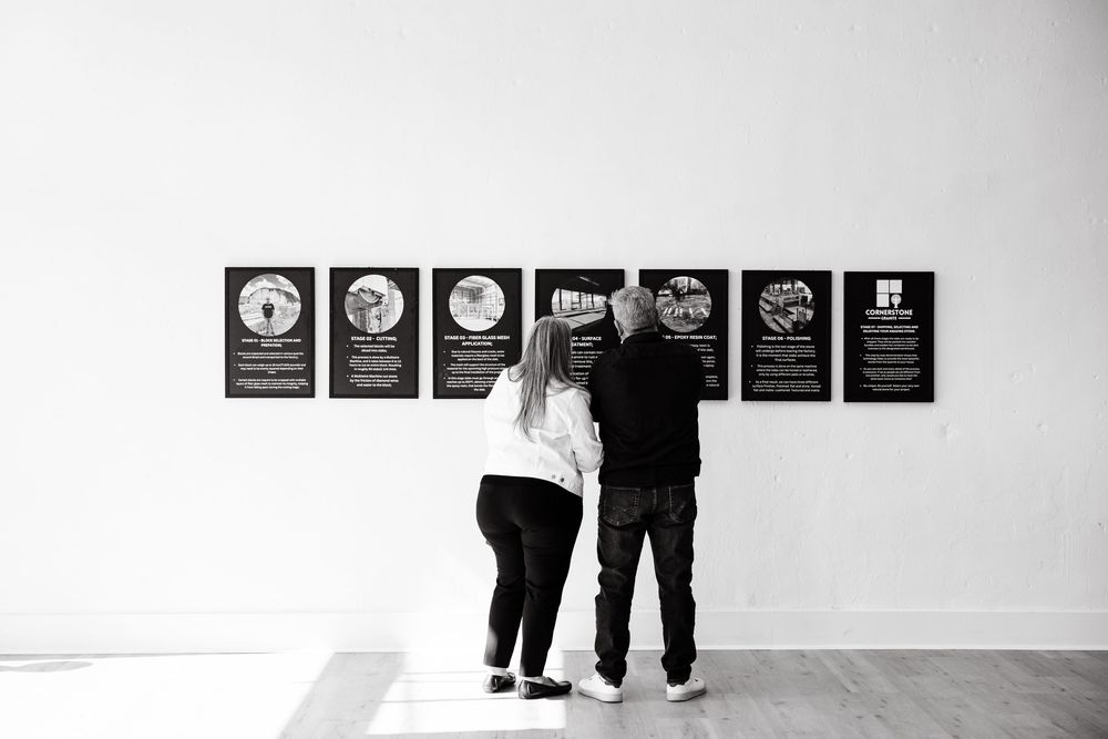 Two people walk past a white wall displaying black and white framed artwork in a gallery setting.