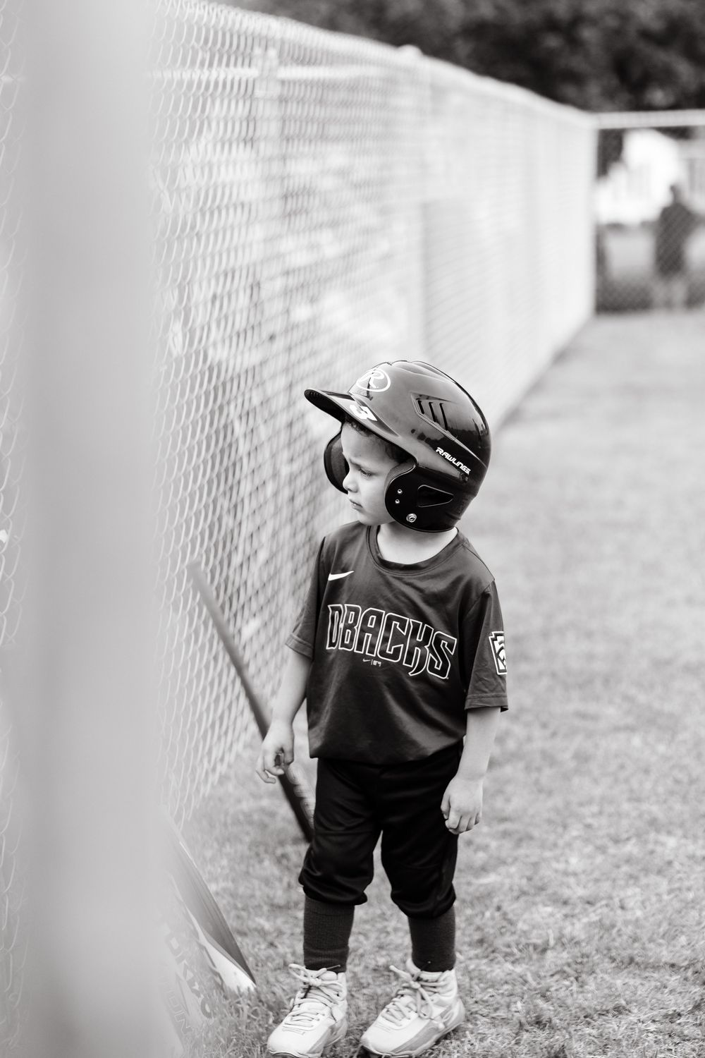 Young boy in Dbacks baseball uniform and helmet standing by chain-link fence in black and white photo.