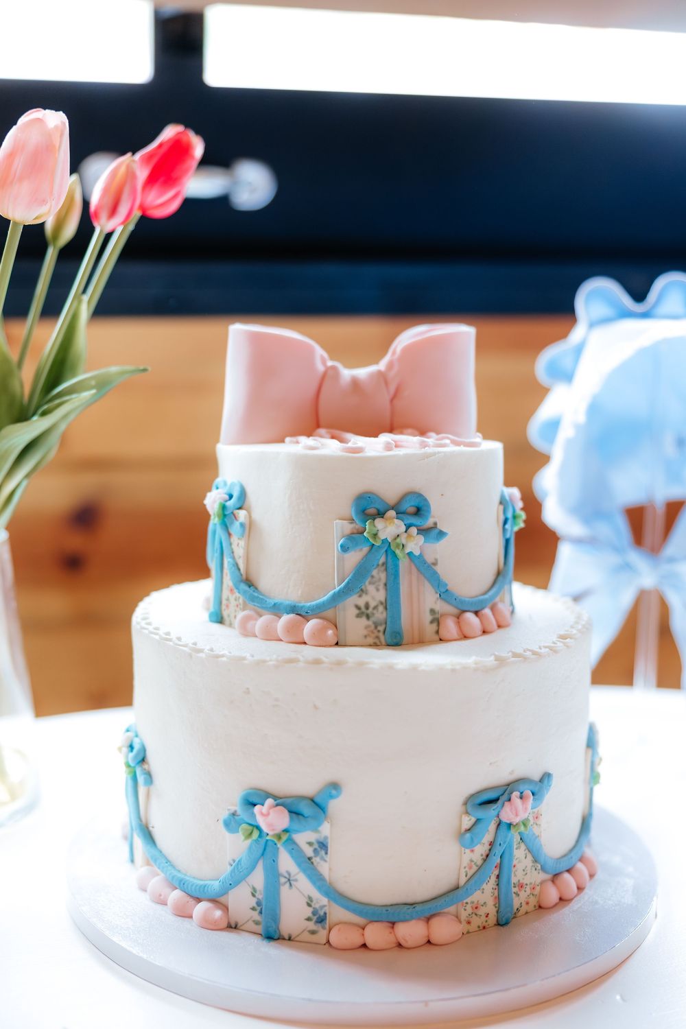Two-tier white cake with blue fondant swags, pink bow topper, and pearl accents for a elegant celebration.