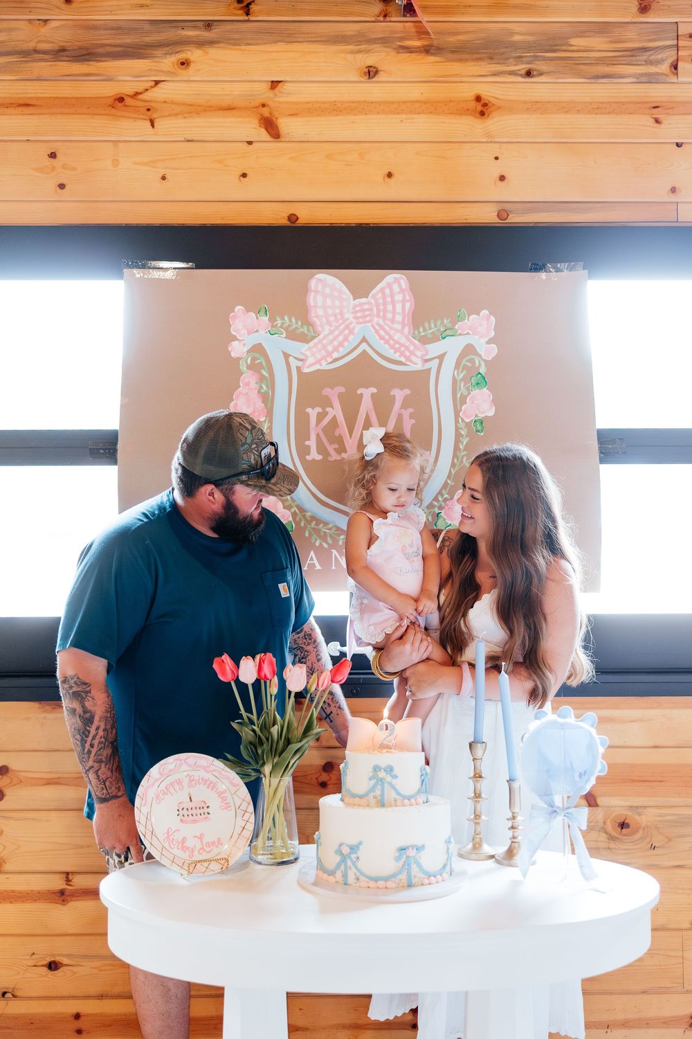 Family celebrating a baby girl's first birthday with a decorated cake and floral monogram backdrop in a rustic wood-paneled venue.