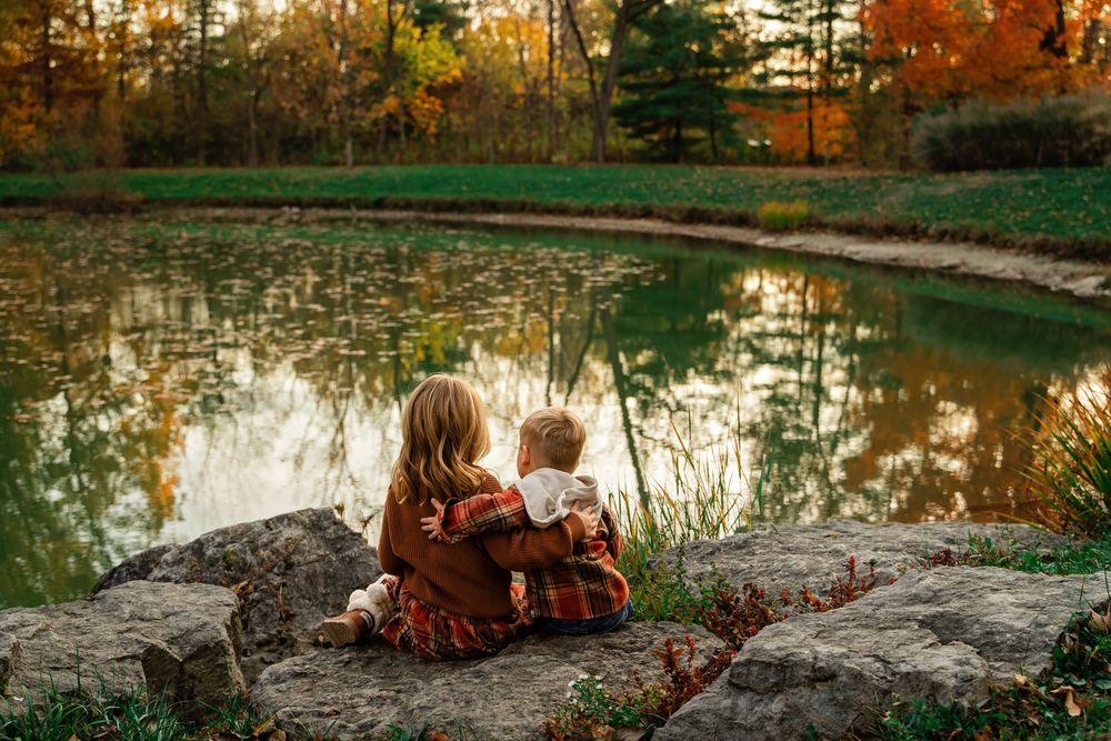 Two young children sitting together on rocks gazing at a peaceful pond reflection in autumn.