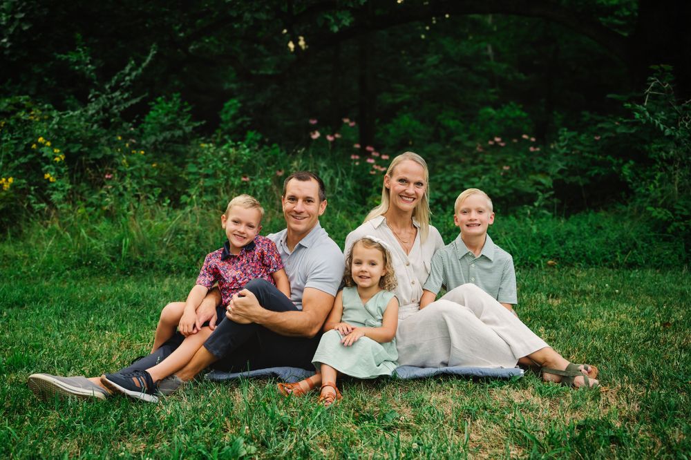 Happy family of five sitting on grass in a lush green park for an outdoor family portrait session.