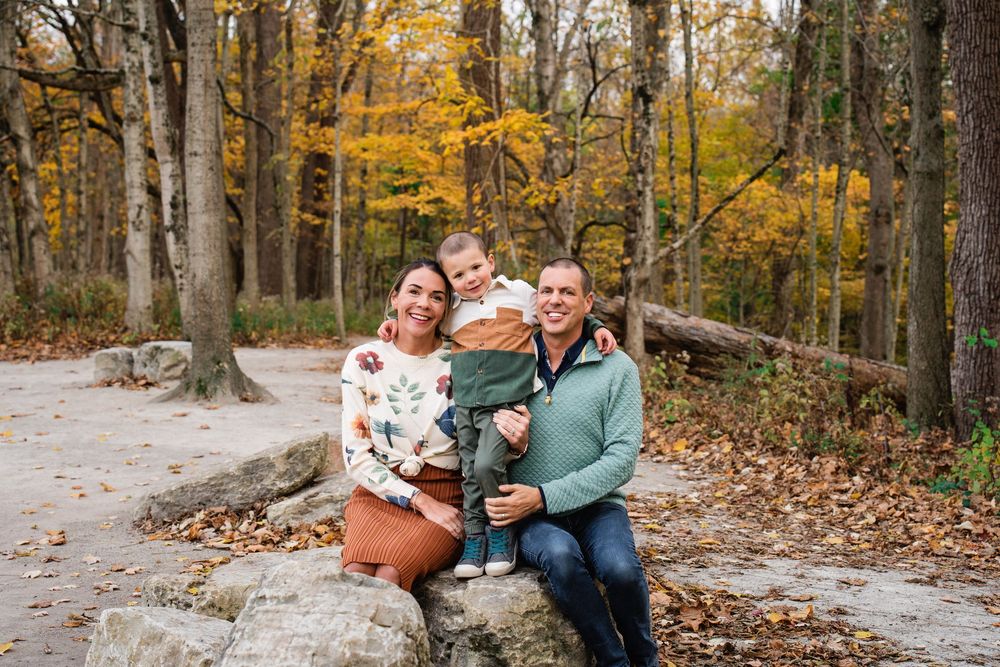 Smiling family of three sitting on rocks surrounded by vibrant fall foliage in the woods.