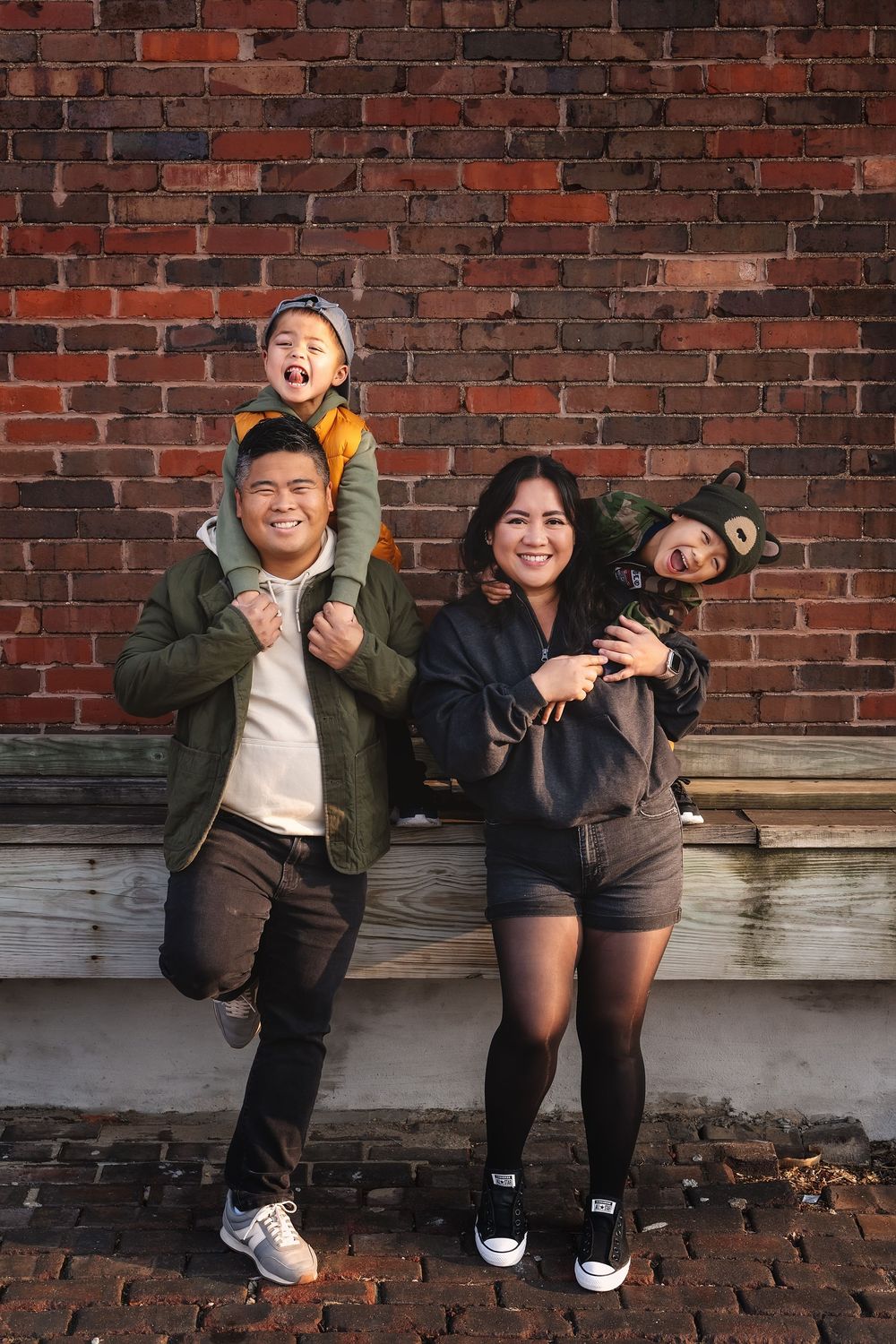 Joyful Family of four posing against brick wall, parents carrying laughing children in Halloween costumes.