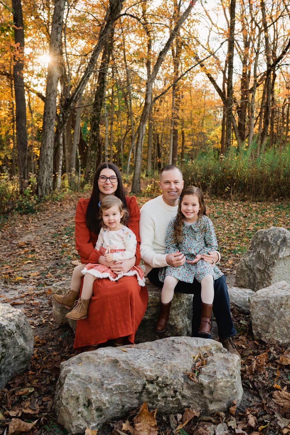 Family of four sitting on rocks in a sunlit autumn forest during a fall photo session.