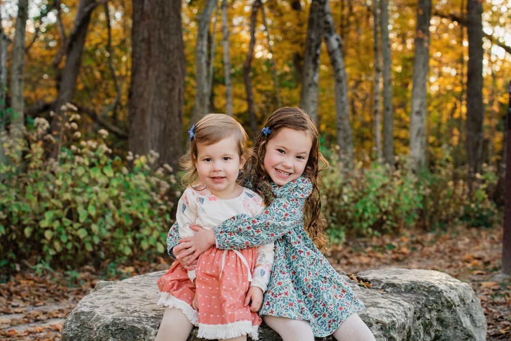 Two young sisters in floral dresses sitting together on rocks in a colorful autumn forest.