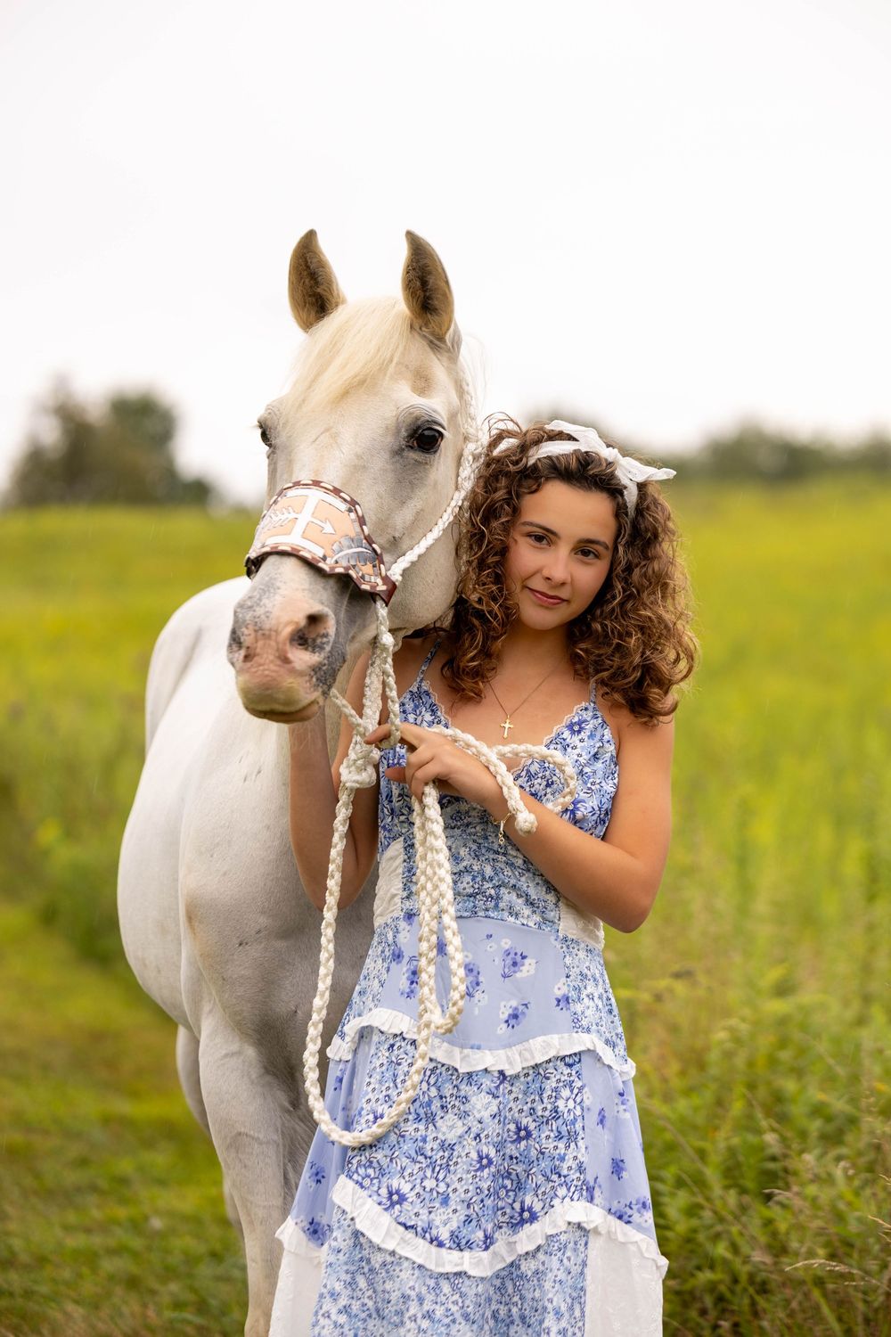 A white horse stands in a green field next to someone wearing a blue and white floral summer dress.
