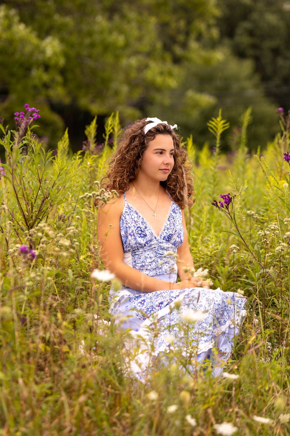 A person in a blue and white floral dress sits thoughtfully among wildflowers in a summer meadow.