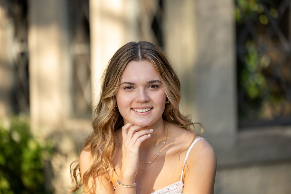 A young woman in a white top smiles outdoors with her hand under her chin in warm sunlight.