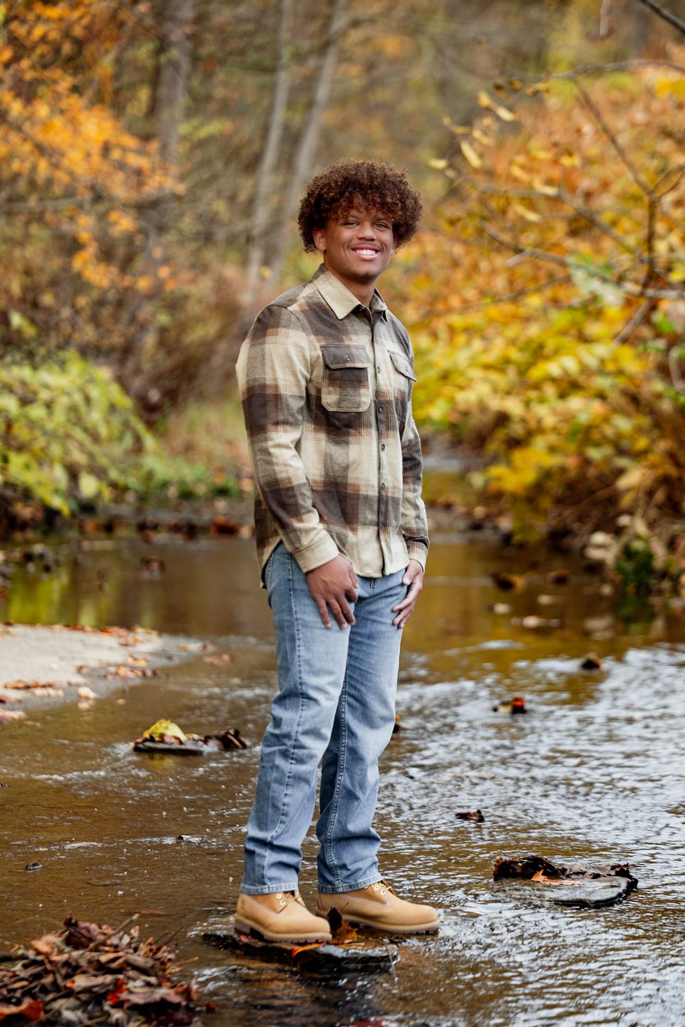 Person in plaid shirt and jeans stands by a scenic autumn creek with golden foliage in the background.