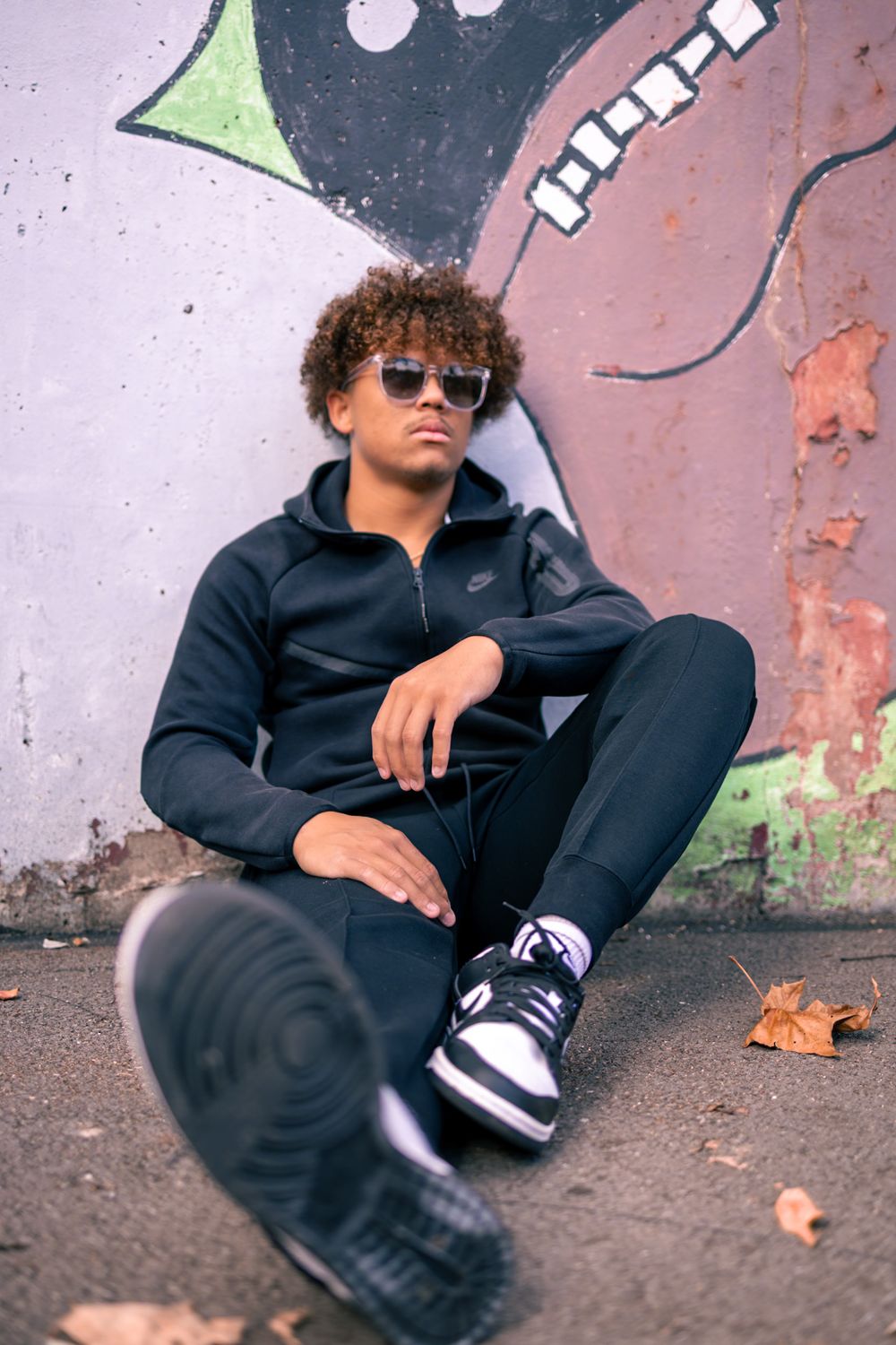Person in black athletic wear relaxes against graffiti wall wearing sunglasses and sneakers.