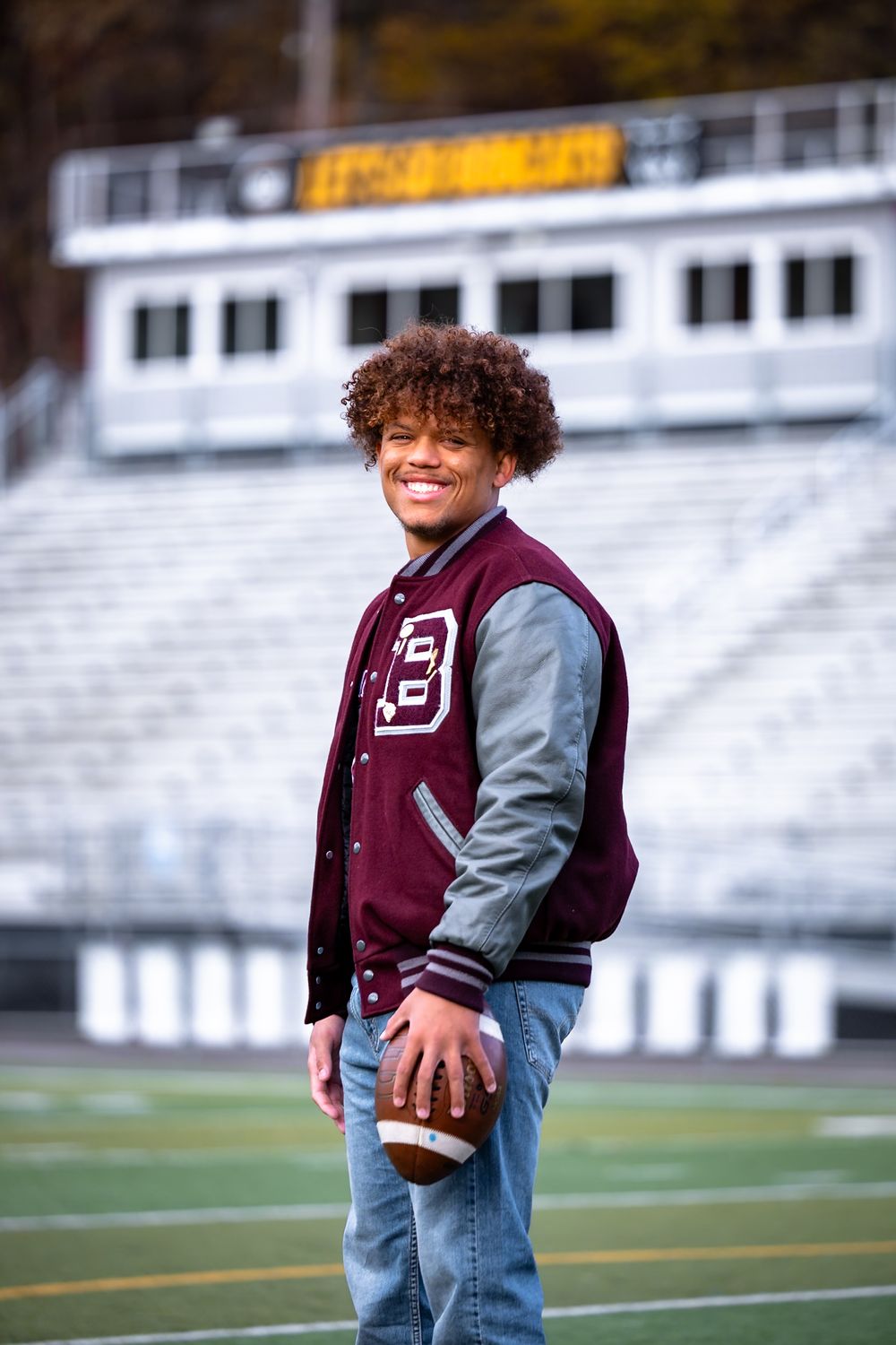 Student athlete in maroon and gray varsity jacket holds football while standing on field with stadium bleachers behind.