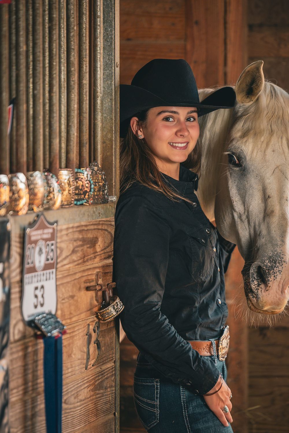 Person in black jacket and hat smiles while petting a white horse in rustic wooden barn setting.