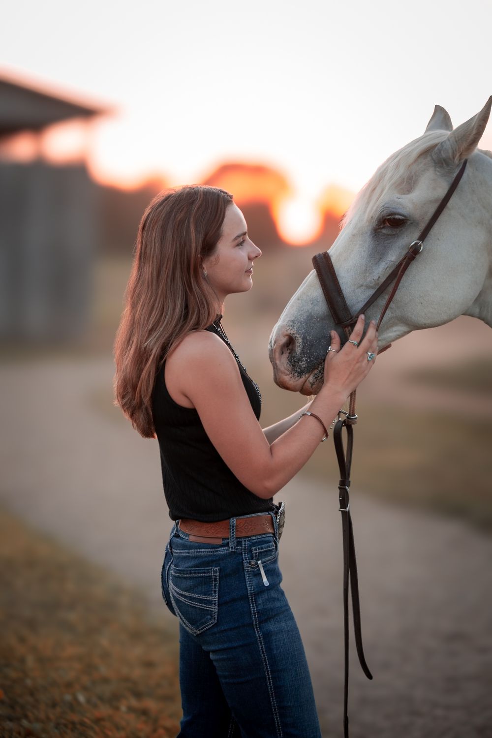 A woman in a black top and jeans shares a tender moment with a white horse at sunset on a dirt path.