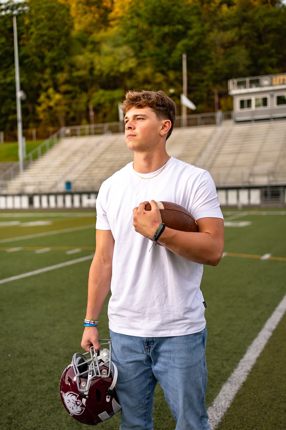 Football player stands on field holding ball and helmet while wearing white t-shirt and jeans during autumn season.